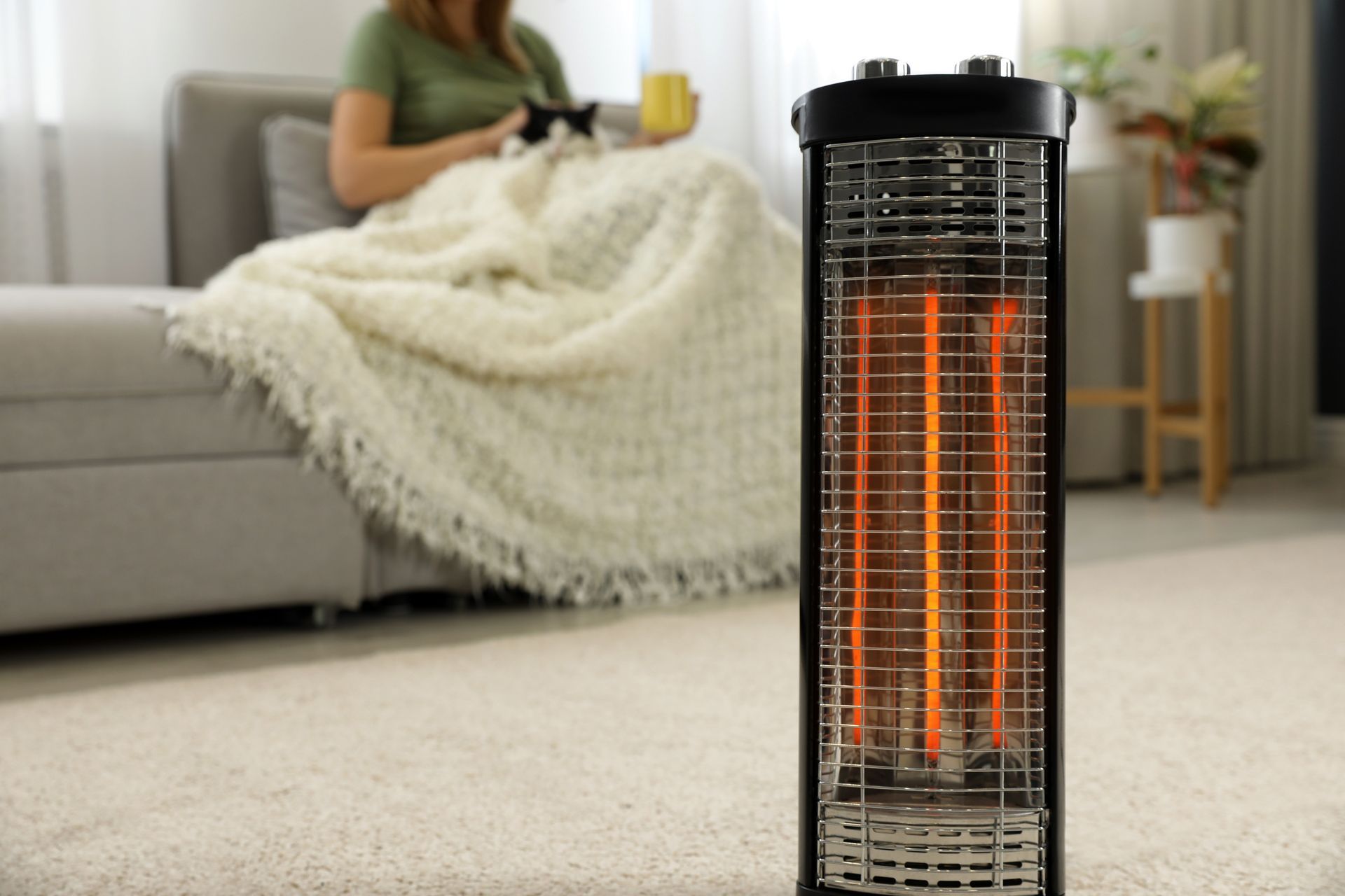 A standing space heater with glowing coils warms a person on a couch.