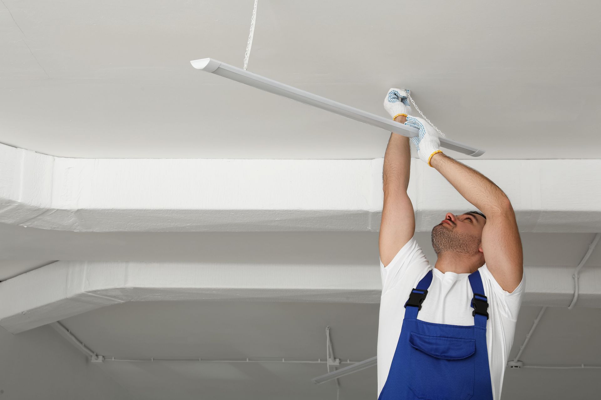 Man in overalls installing a light fixture on a white ceiling.