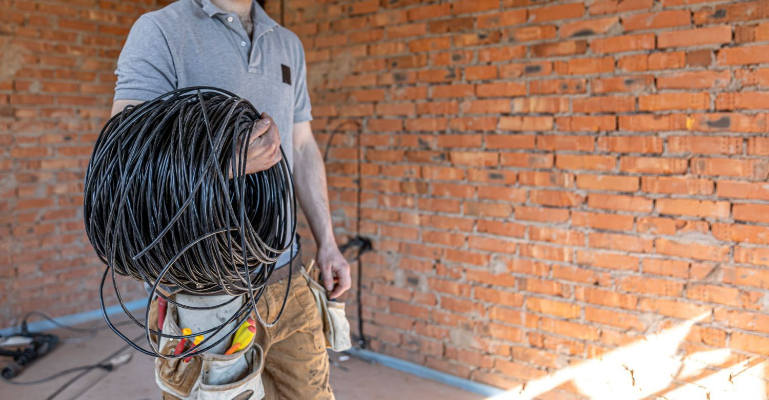 Man holding a coil of black electrical wire in a room with a brick wall, wearing work clothes.
