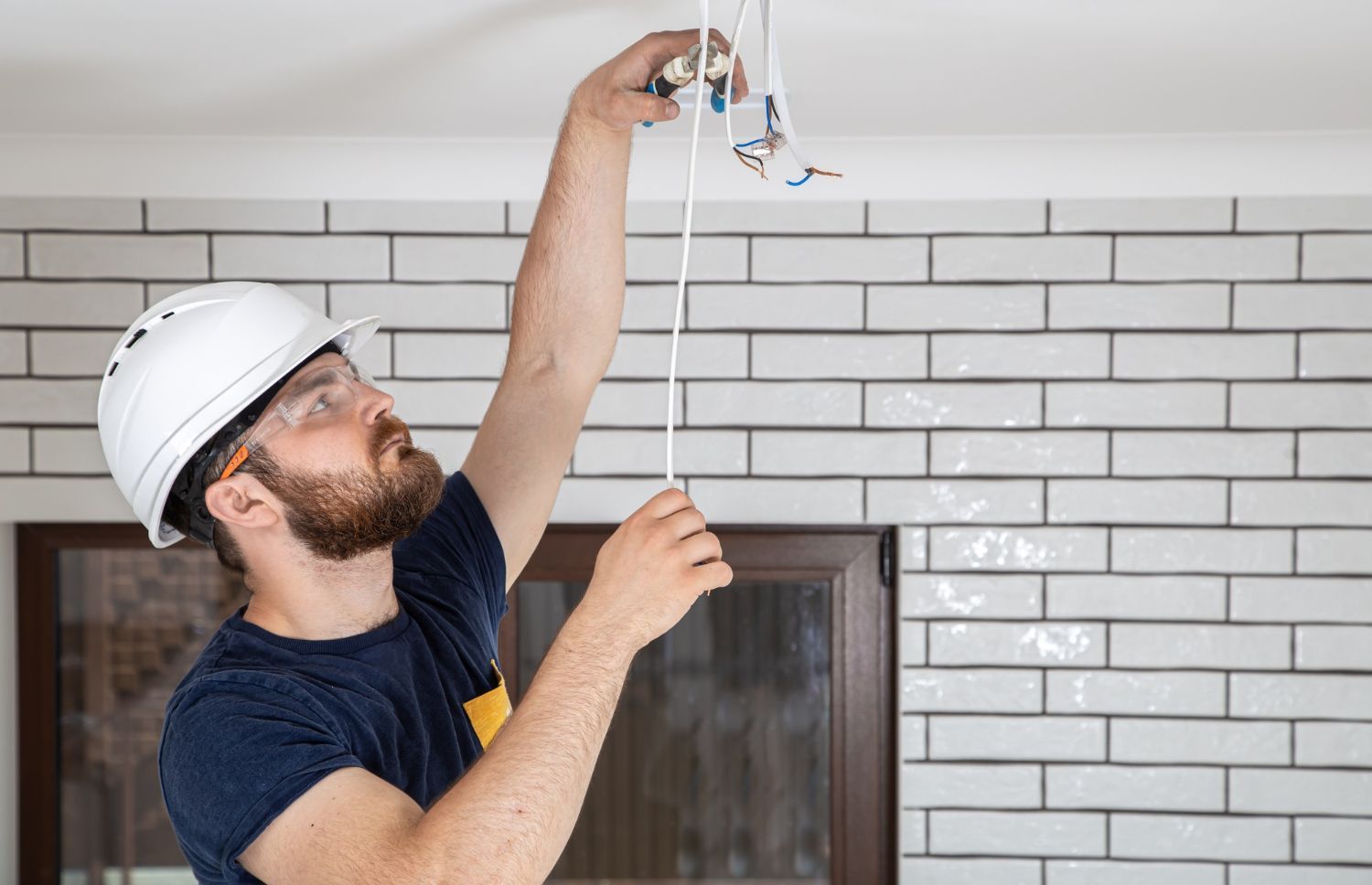 Electrician in a white hard hat and safety glasses working on wires, indoors.