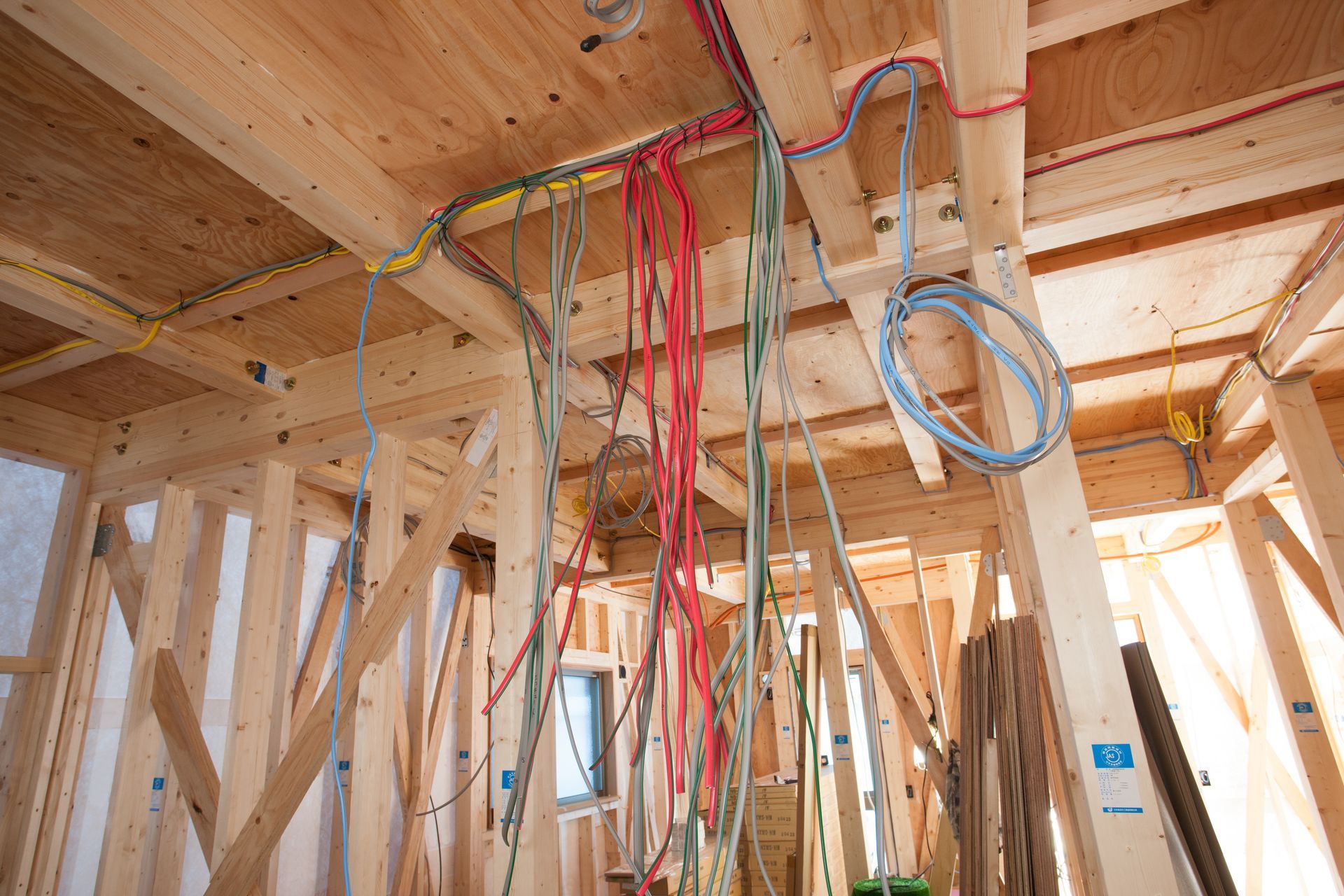 Exposed wooden house framing with hanging red, blue, and yellow electrical wires during construction