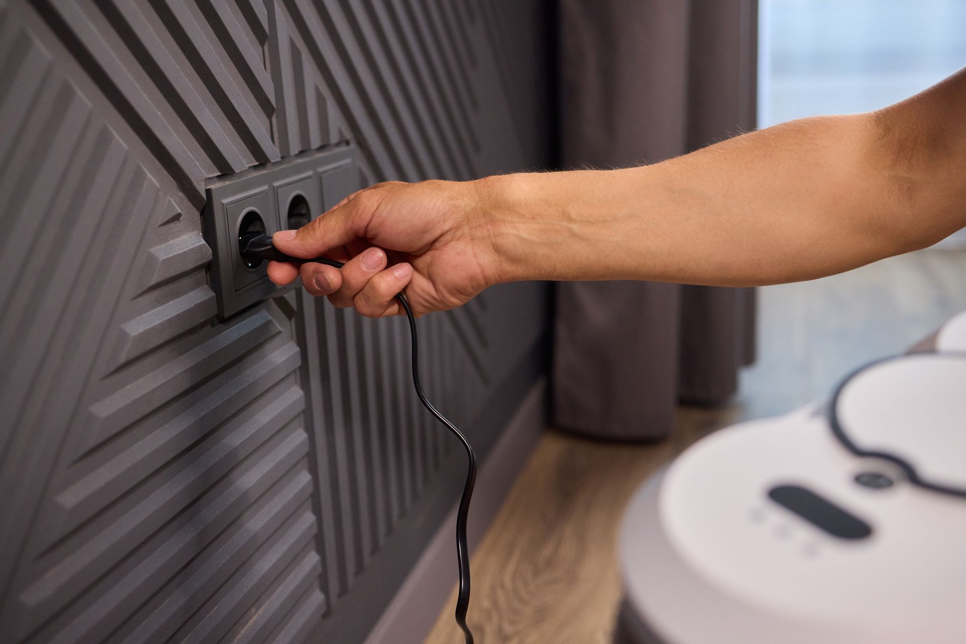 Person plugging a black cord into a dark gray wall outlet, near a white robot vacuum.