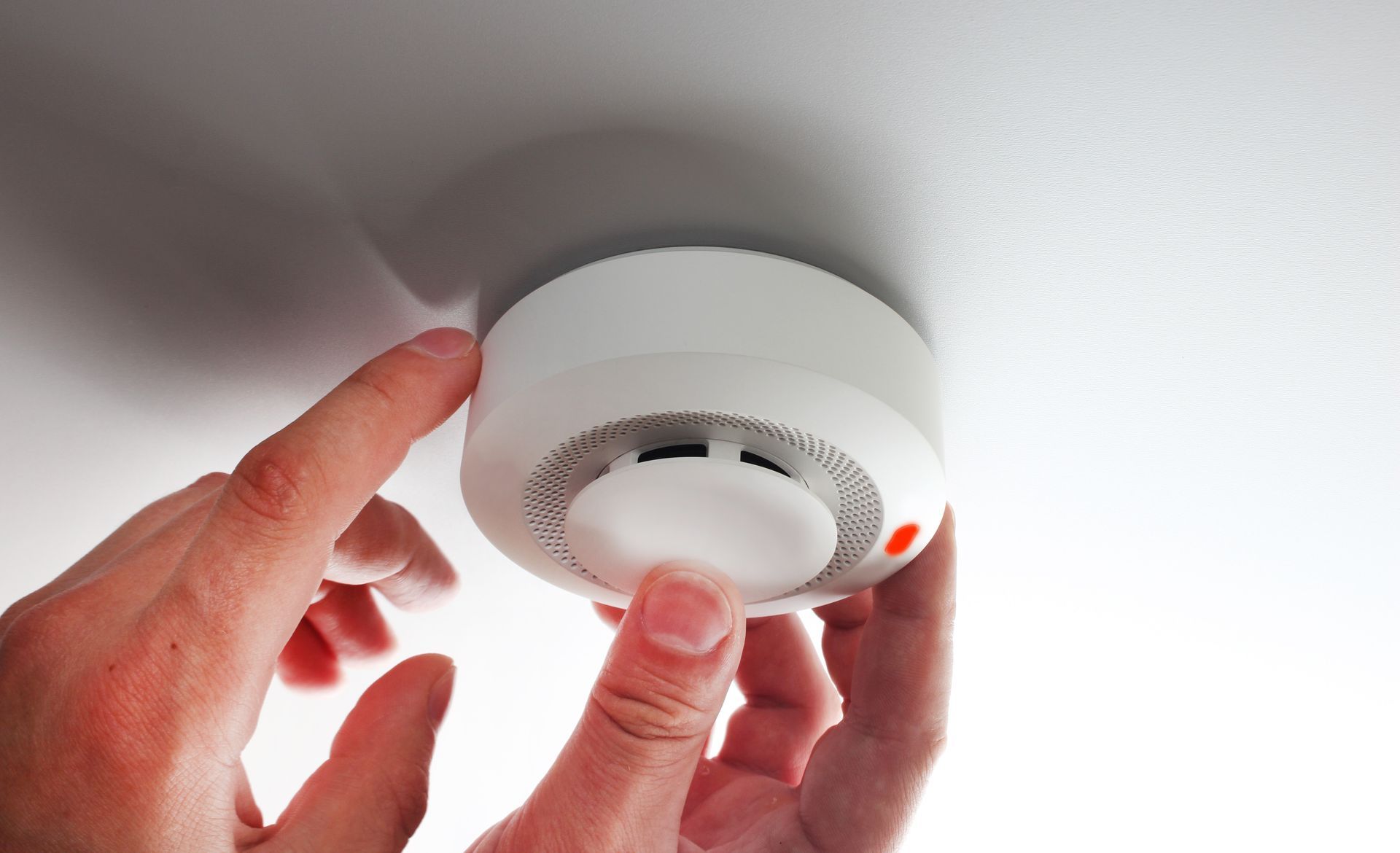 Hands installing a white smoke detector on a white ceiling.