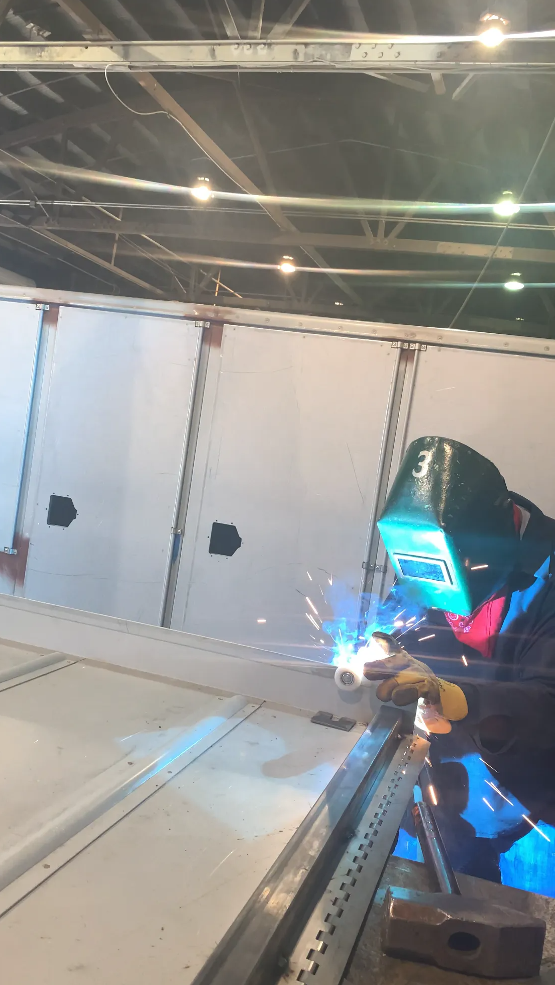 Welder in a workshop, wearing a helmet, welding metal with sparks and blue light.
