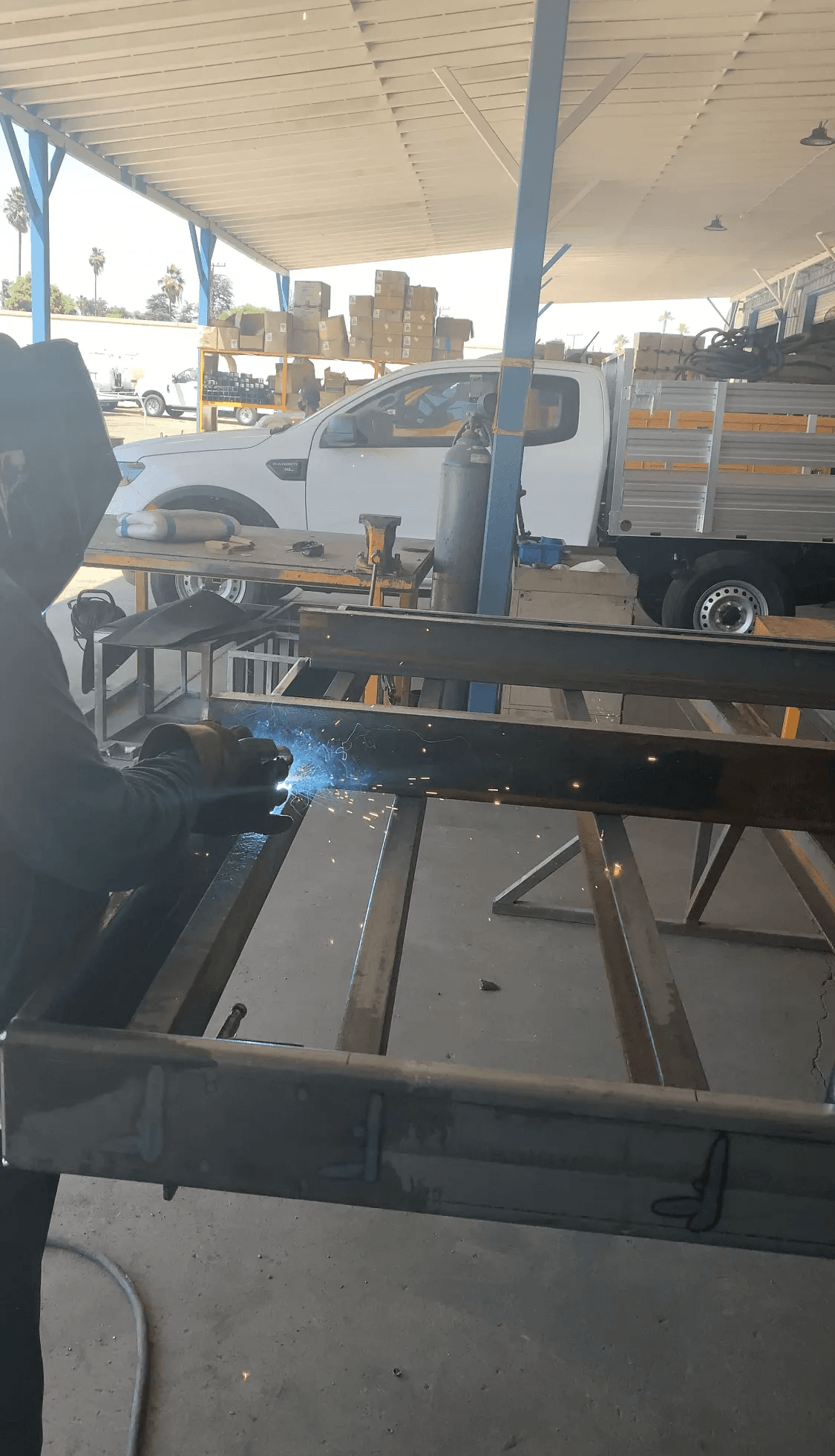 A person welds metal in a workshop, sparks flying. A white truck and building are in the background.
