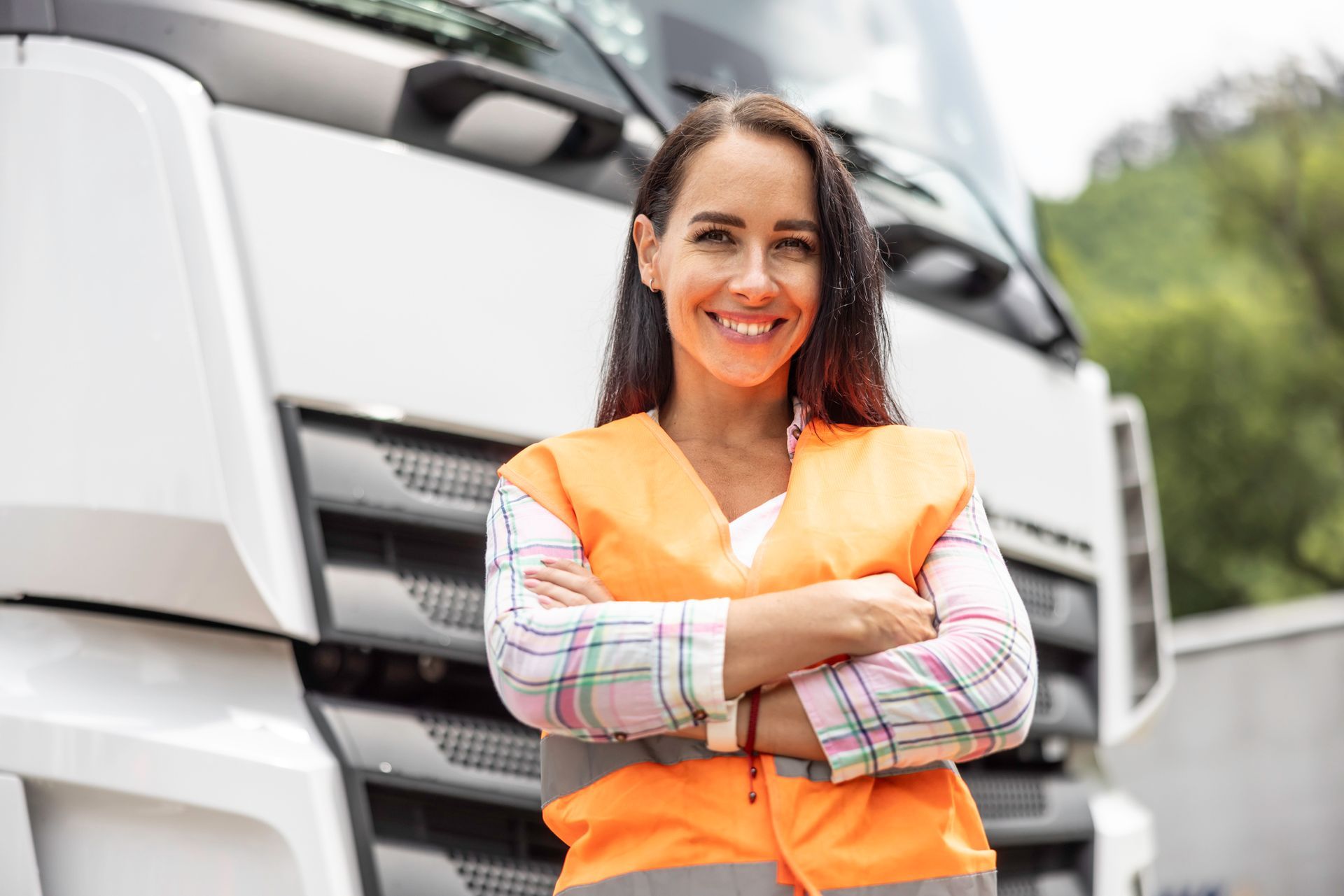 Smiling woman with arms crossed wears orange safety vest, standing in front of a semi truck