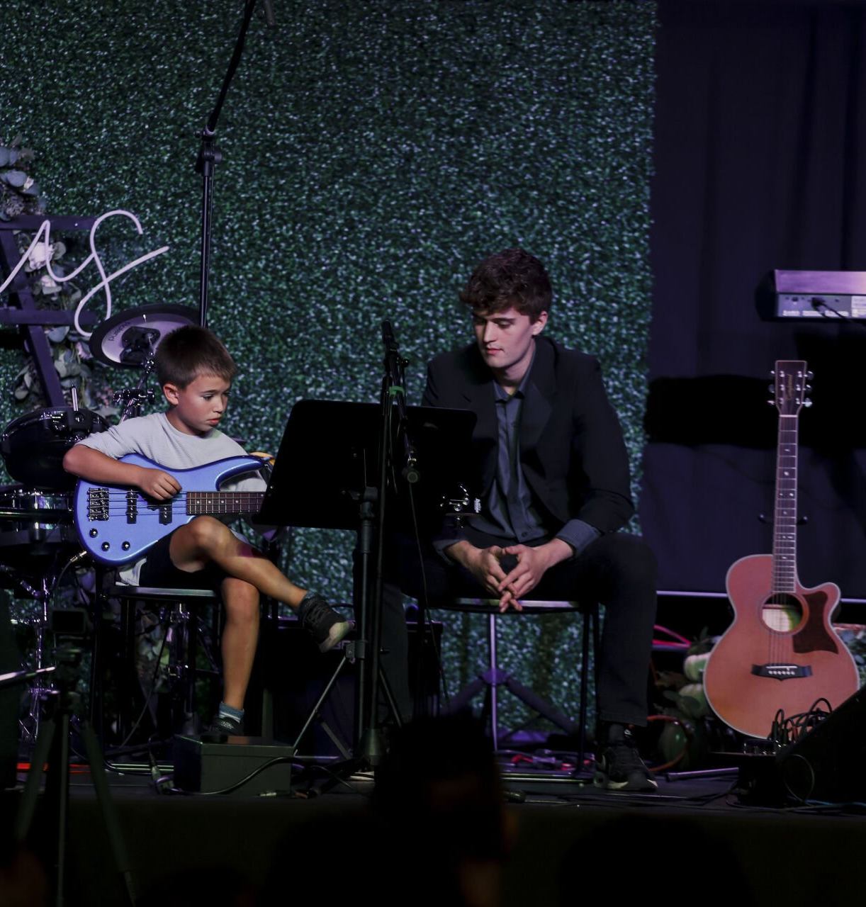 A young man is supporting a young boy play a guitar on stage.