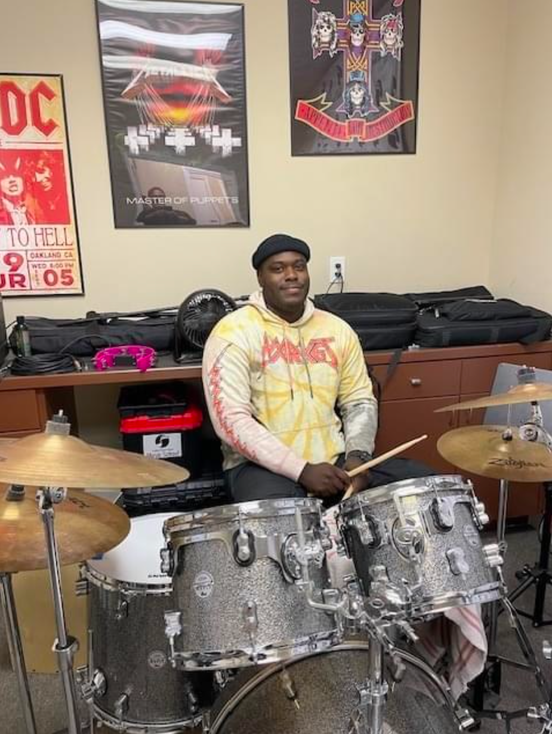 A man is playing drums in a room with posters on the wall.