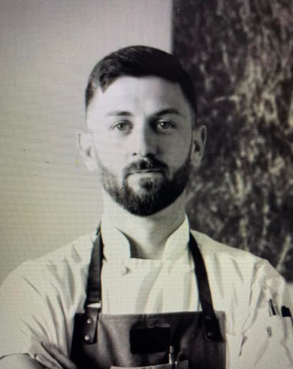 A man with a beard is wearing an apron and a chef 's jacket.