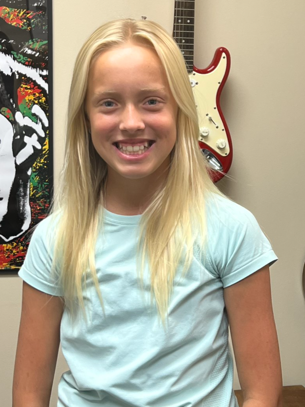 A young girl (student) is smiling in front of a guitar