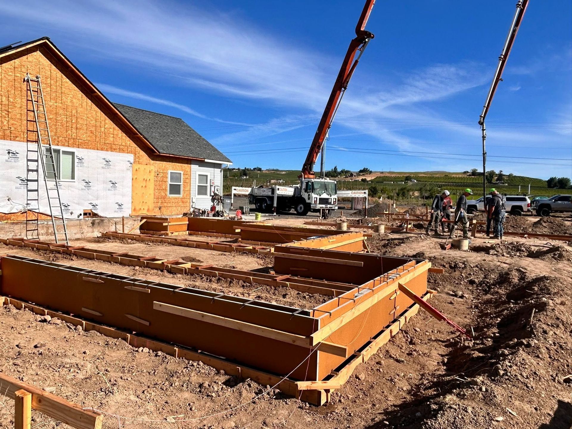 Construction site with concrete foundation forms, crane, workers, and a house under construction under a blue sky