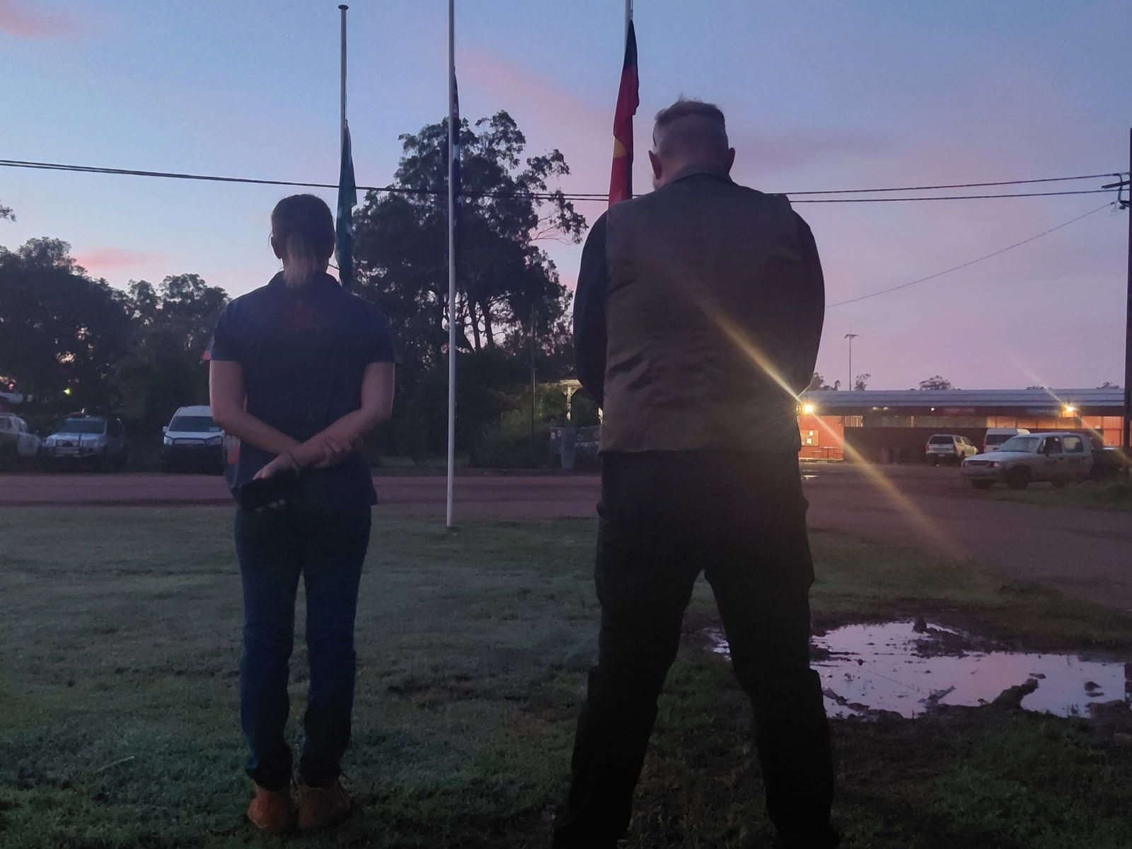 Two people standing with their back to the camera. They look at the four flag poles as the sun rises. 