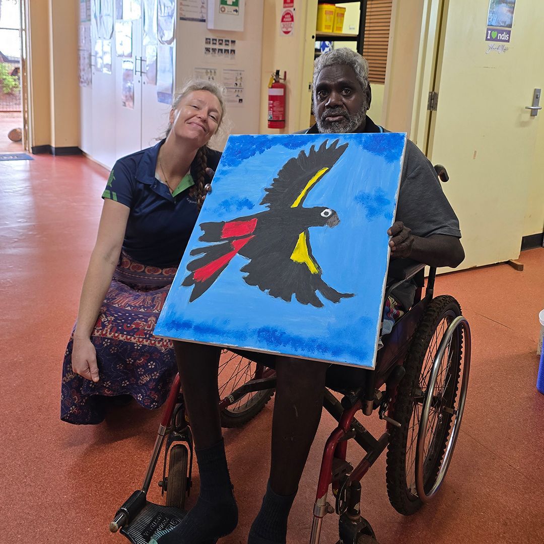 A man in a wheelchair holds a painting of a black cockatoo. A woman is next to him. 