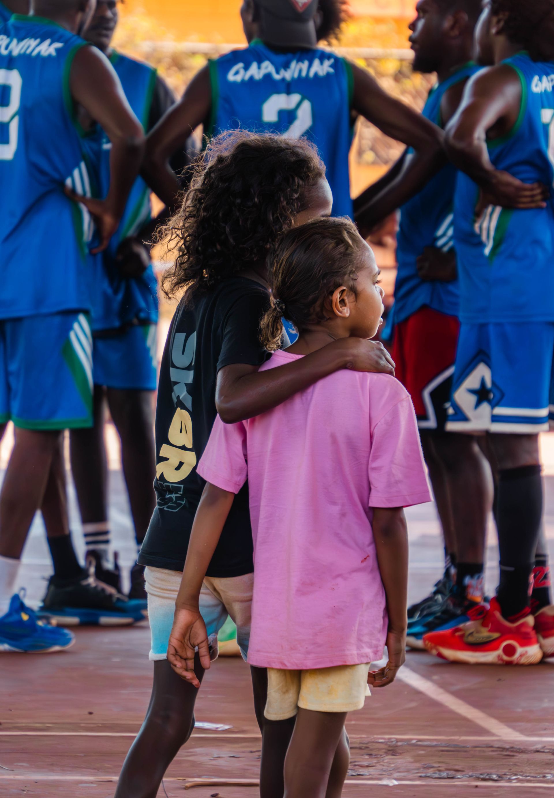 Two children with their arms around each other as they watch a basketball team prepare for a game.