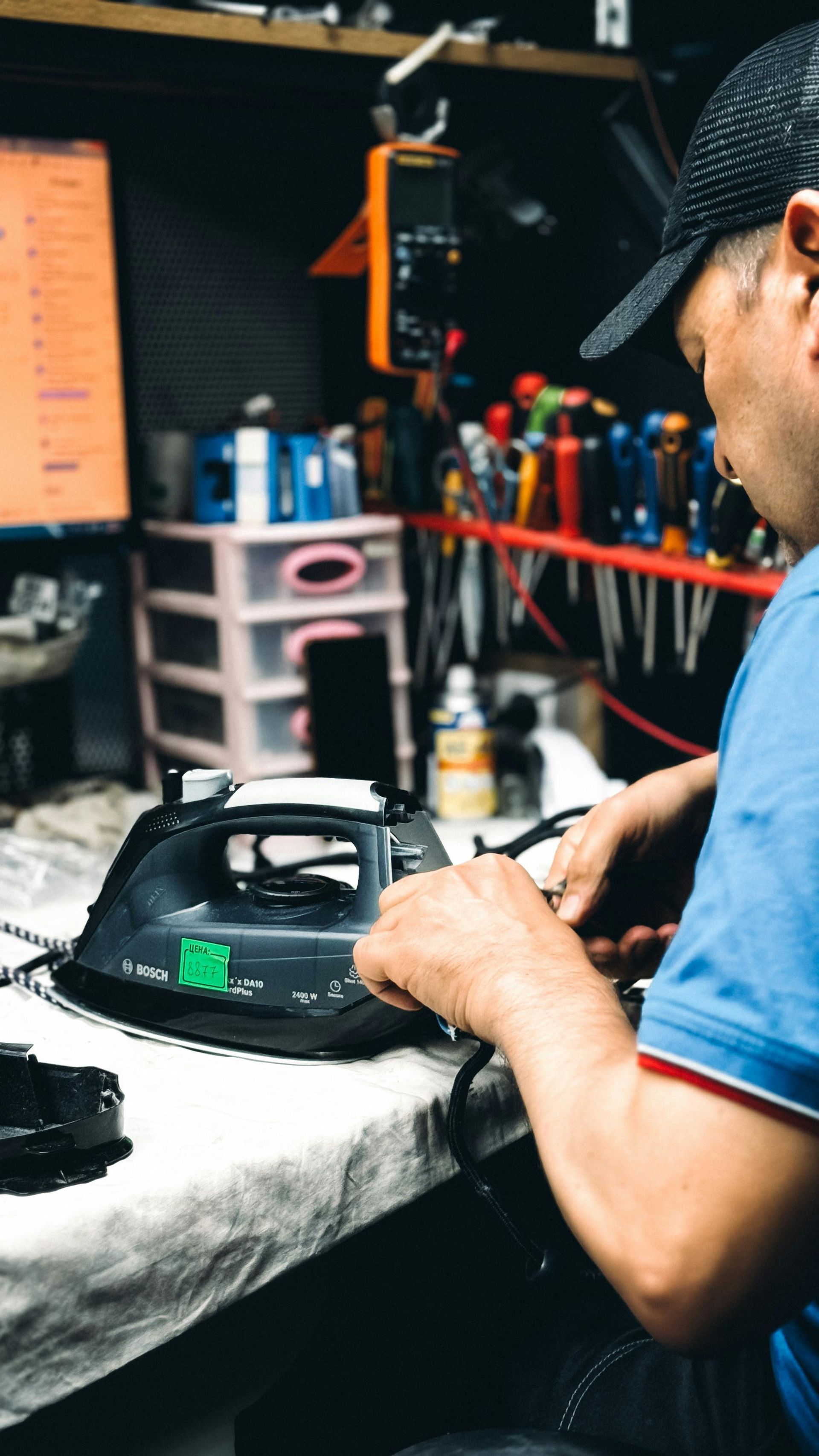 A technician repairs a black steam iron on a workbench surrounded by tools in a workshop.
