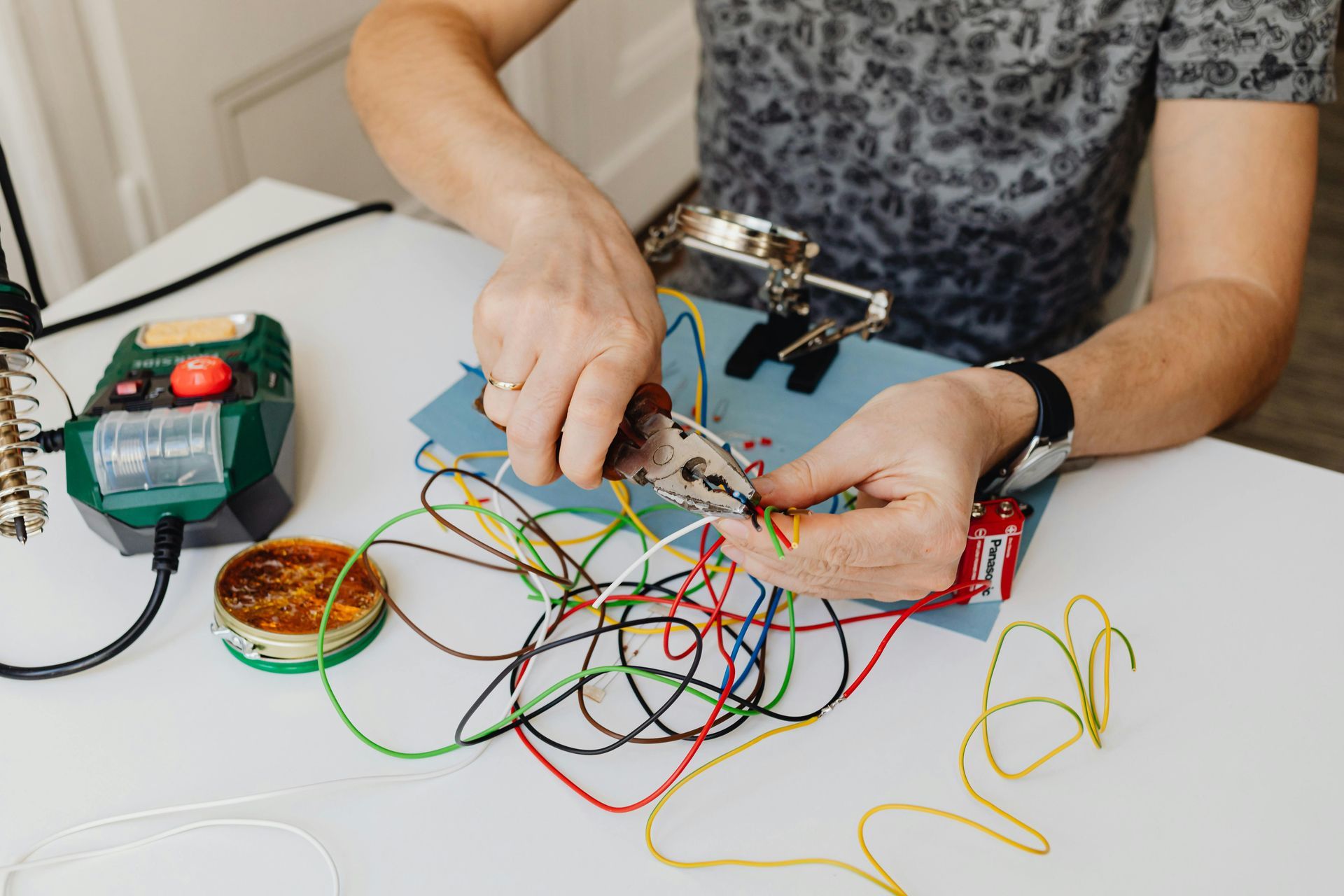 Hands using pliers to work on colored electrical wires at a desk with a soldering station and supplies.