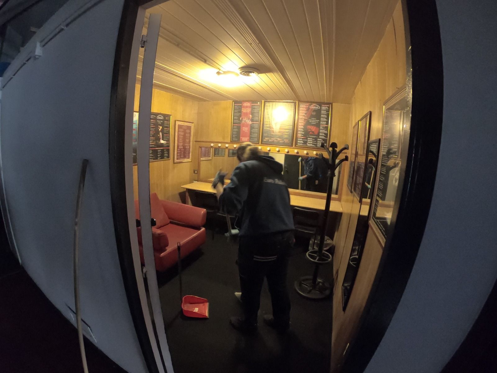 Una persona in piedi all'interno di una piccola stanza rivestita in legno. La stanza è arredata con poster, un divano rosso e una chitarra.