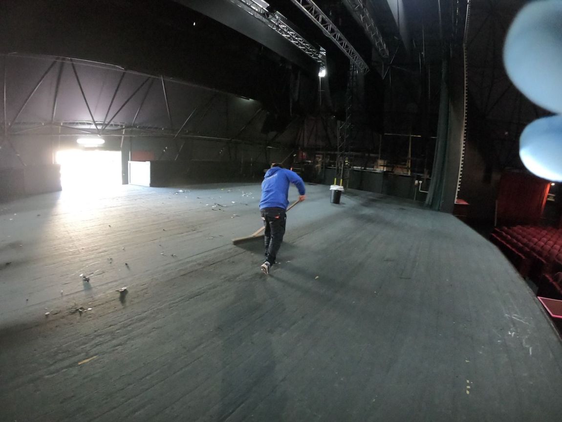 Una persona spazza un grande palcoscenico grigio in un teatro. Le porte posteriori sono aperte, lasciando entrare la luce.