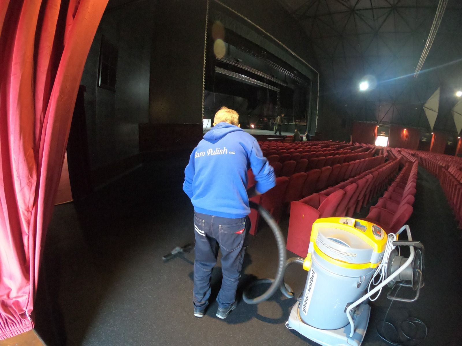 Persona che passa l'aspirapolvere nella zona dei posti a sedere di un teatro. Indossa una felpa blu con cappuccio e guarda dall'altra parte. Sedili e tende rossi.
