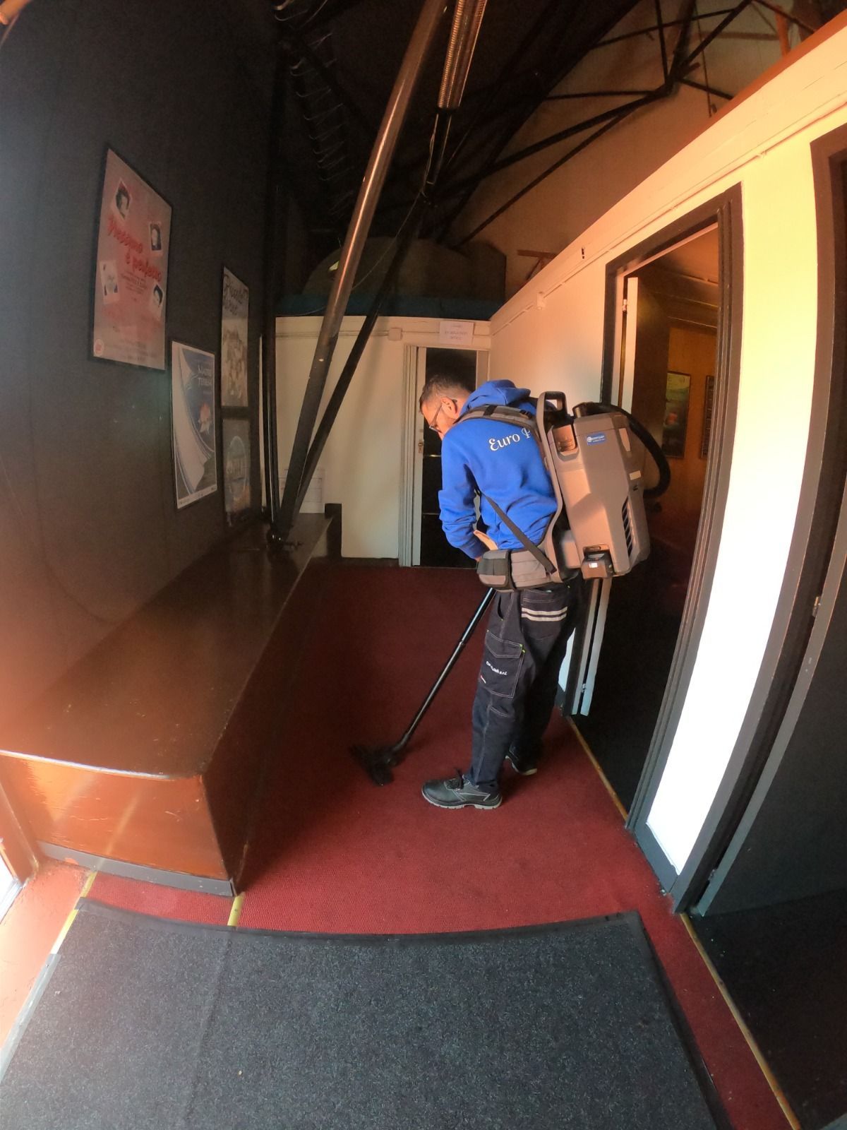 Persona che passa l'aspirapolvere in un lungo corridoio con moquette rossa e quadri alle pareti.