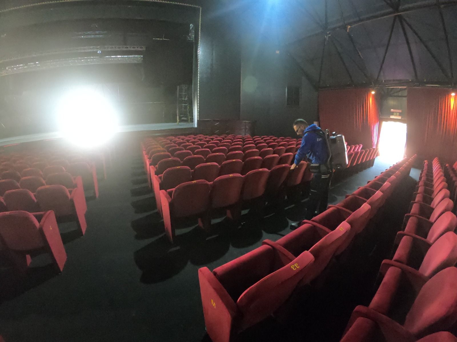 File di poltrone rosse da teatro, palcoscenico illuminato a giorno, persone al lavoro.