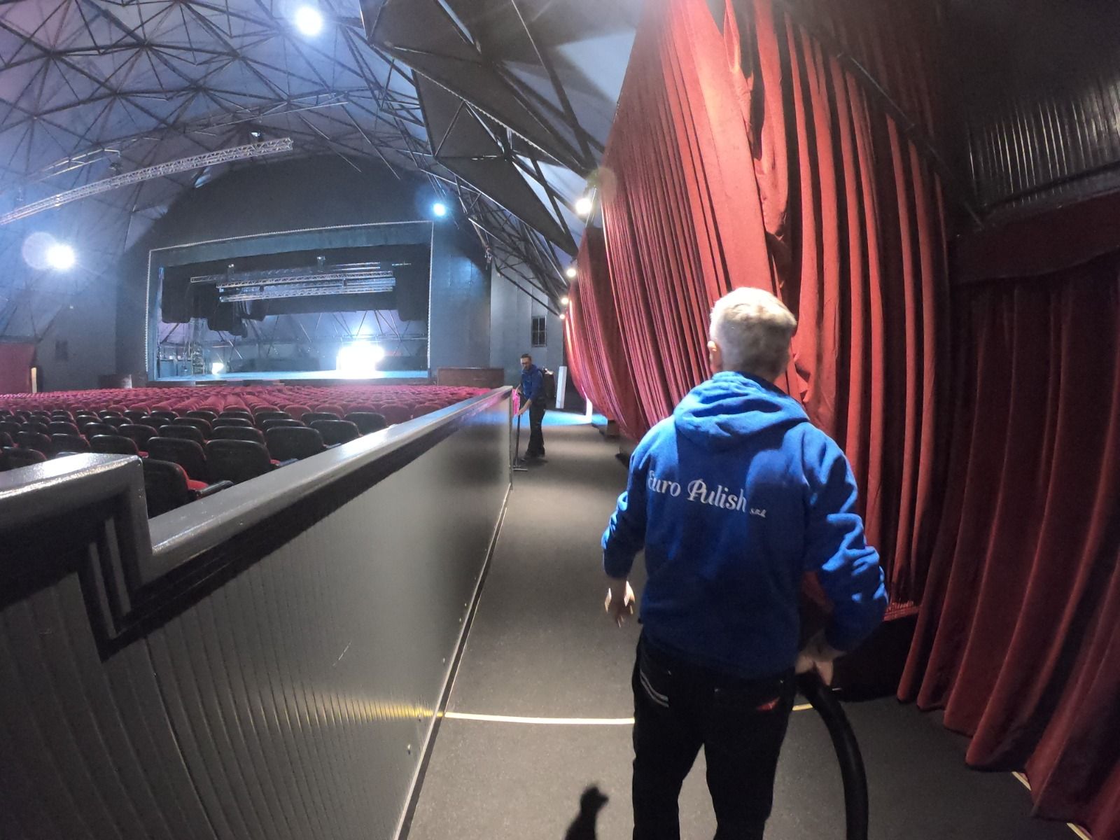 Un uomo con una felpa blu cammina dietro le quinte di un teatro, con le tende rosse sulla destra e il palco sullo sfondo.