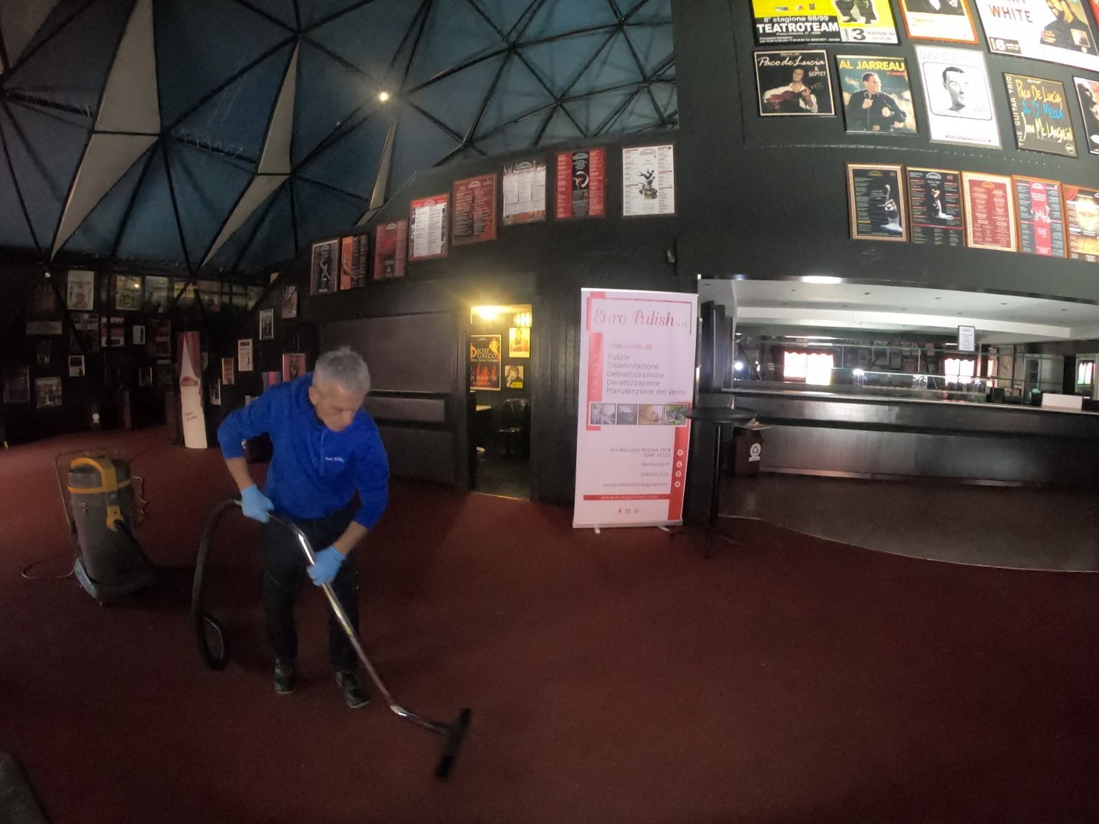 Persona che passa l'aspirapolvere sul tappeto rosso nella hall di un teatro con poster alle pareti e soffitto a cupola.