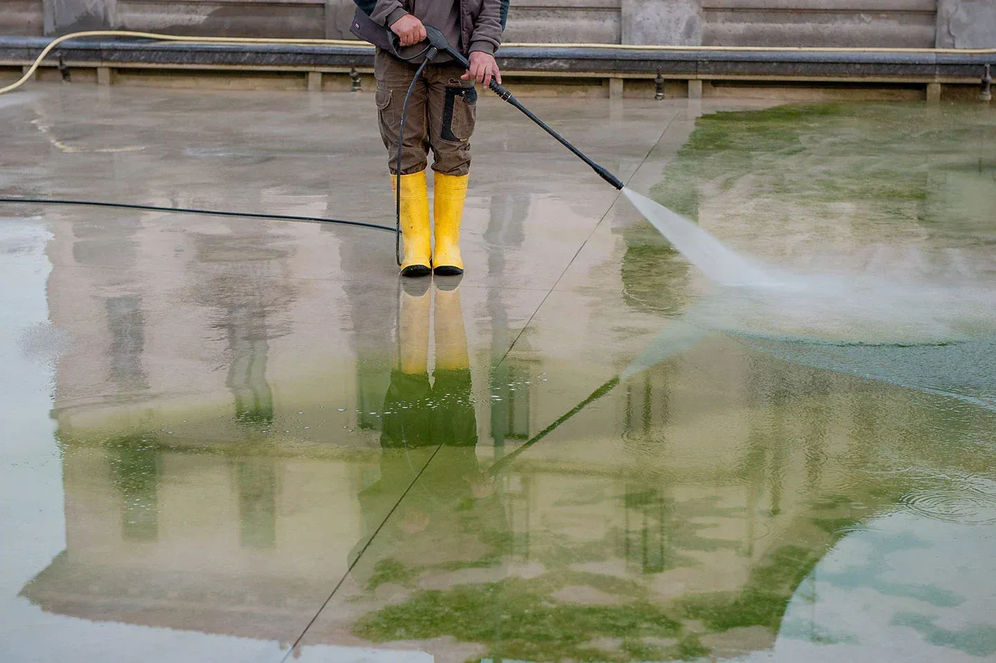 A man in yellow boots is using a high pressure washer to clean a pool.