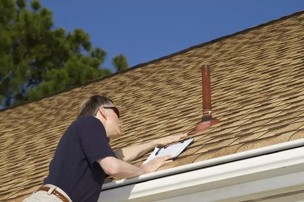 A man is standing on the roof of a house holding a clipboard.