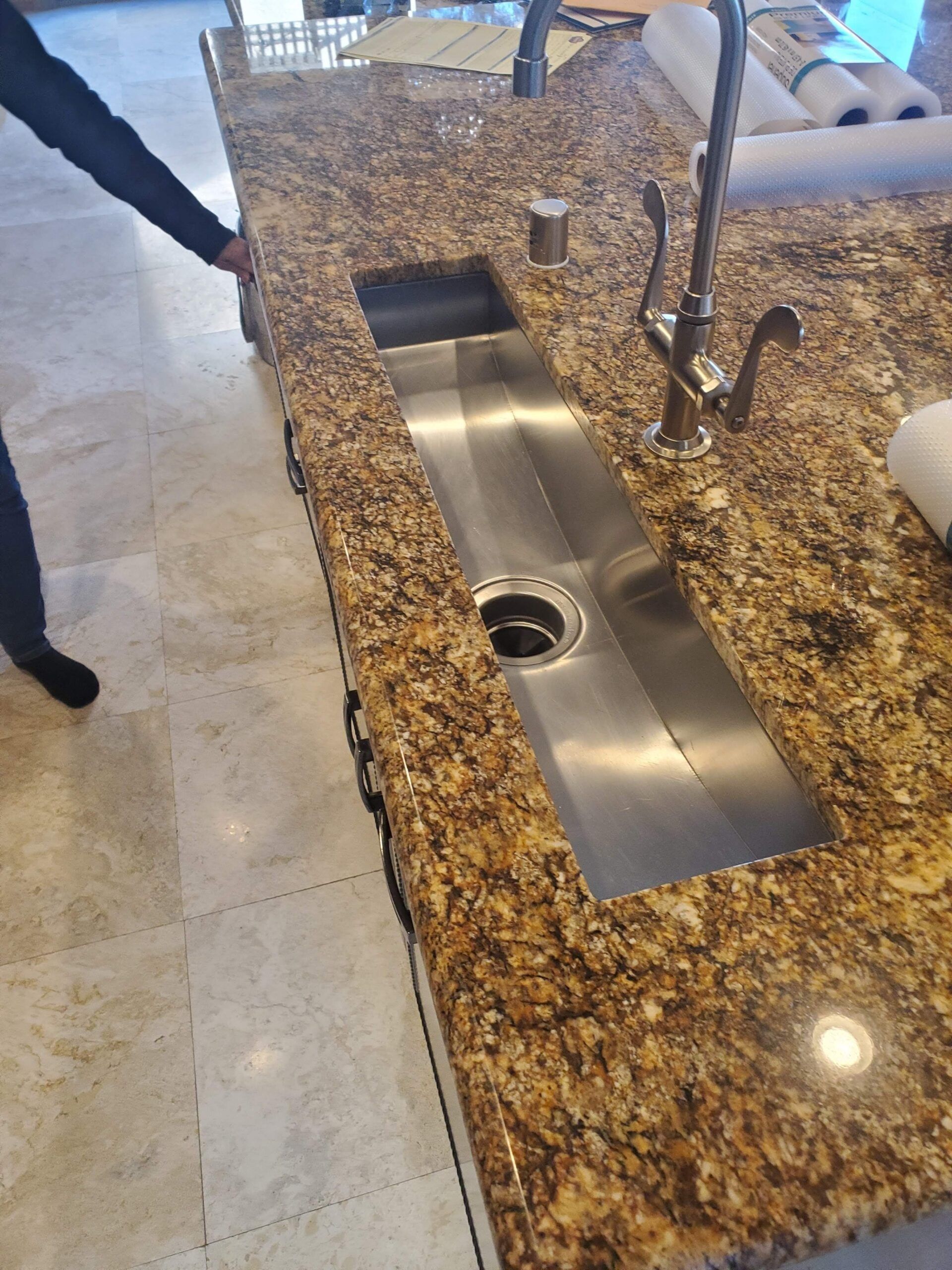 A person is standing next to a kitchen sink with a granite counter top.