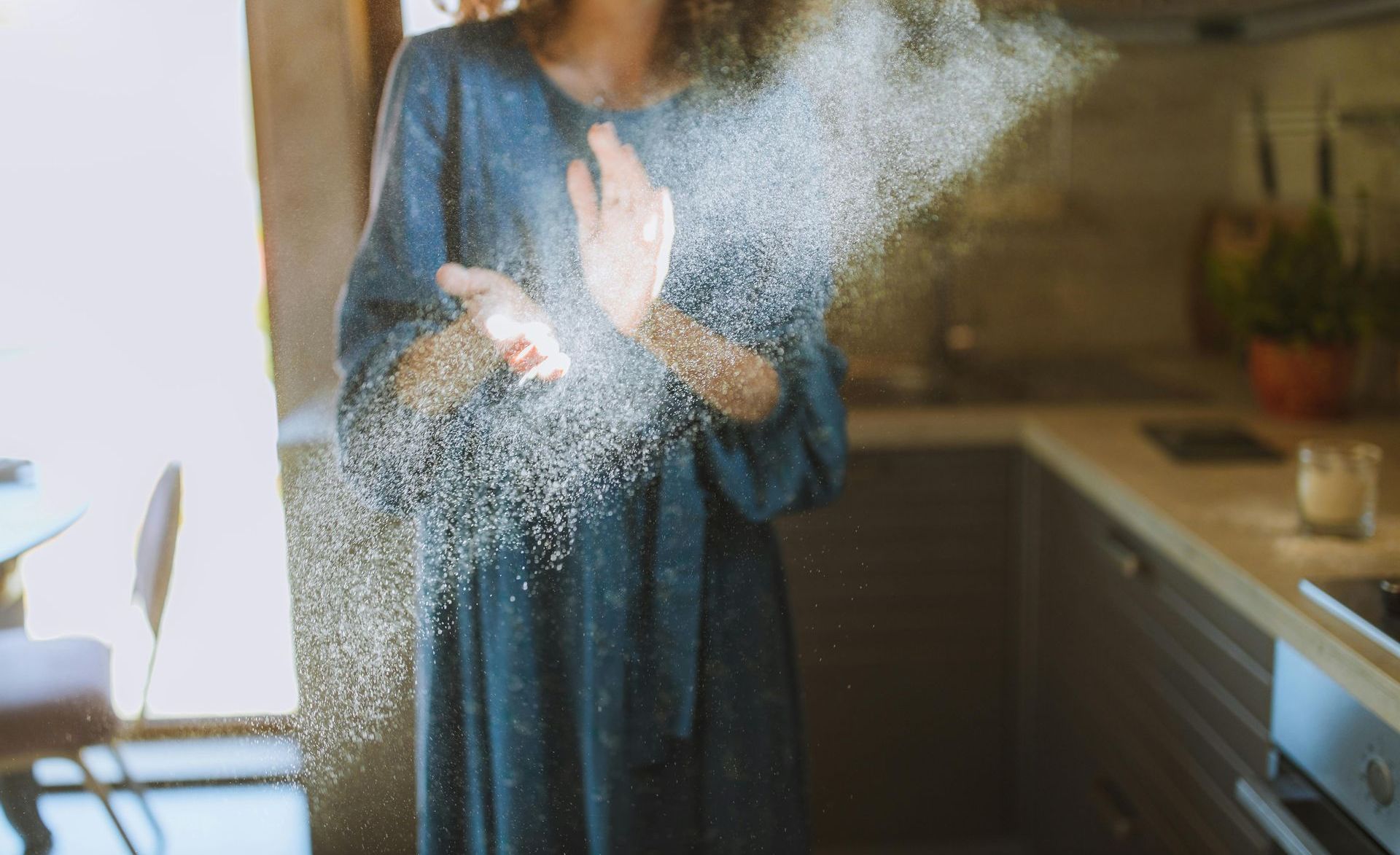 A woman in a blue dress is throwing flour in the air in a kitchen.