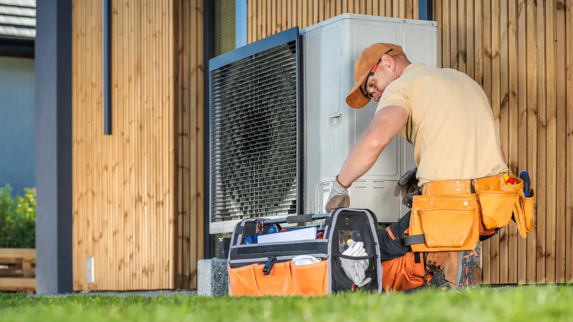 A repairman in an orange hat and tool belt services an outdoor air conditioning unit near a wooden wall.