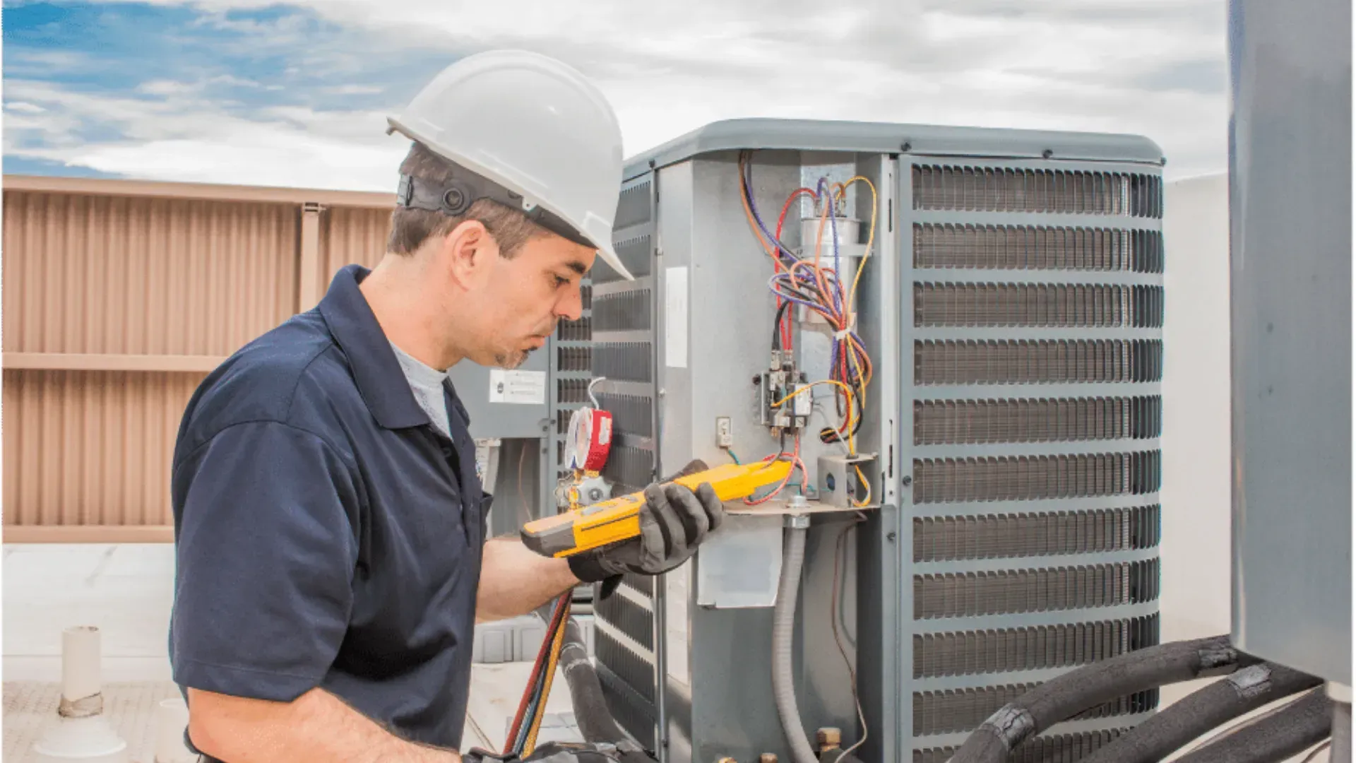HVAC technician in a hard hat, inspecting an air conditioning unit with a multimeter. The technician is outdoors on a rooftop.