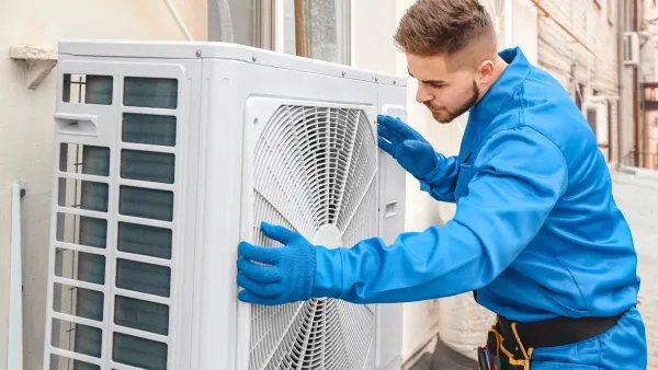 A man in blue work clothes inspects an outdoor air conditioning unit. He wears gloves and is outside.