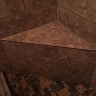 A corner shower seat made of brown, textured square tiles, positioned against two tiled shower walls.