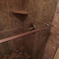 A walk-in shower with brown stone-look wall tiles, a corner shelf, and a metallic shower handle.