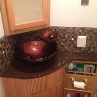 A brown glass vessel sink sits on a dark countertop with a mosaic tile backsplash in a bathroom with wooden cabinetry.
