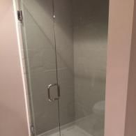 A modern glass shower door with a polished chrome handle, mounted in a neutral-toned bathroom.