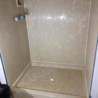 A beige, walk-in shower stall with a floor drain and a light-colored, patterned surround in a bathroom.