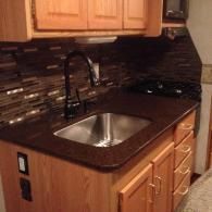 A kitchen counter with a stainless steel sink, dark faucet, and mosaic tile backsplash beneath light wood cabinetry.