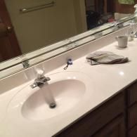 A light-colored bathroom countertop with a sink, faucet, a folded towel, and a small cup, reflecting in a wall mirror.