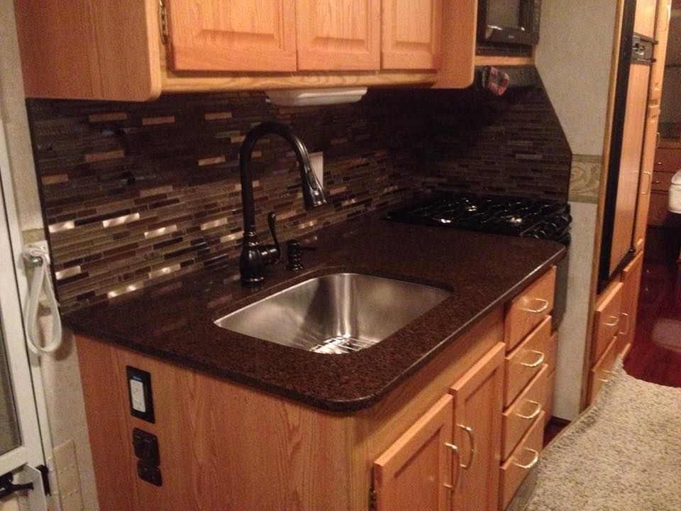 A kitchen in an RV featuring wood cabinets, a dark granite-style countertop, a stainless sink, and a dark mosaic backsplash.