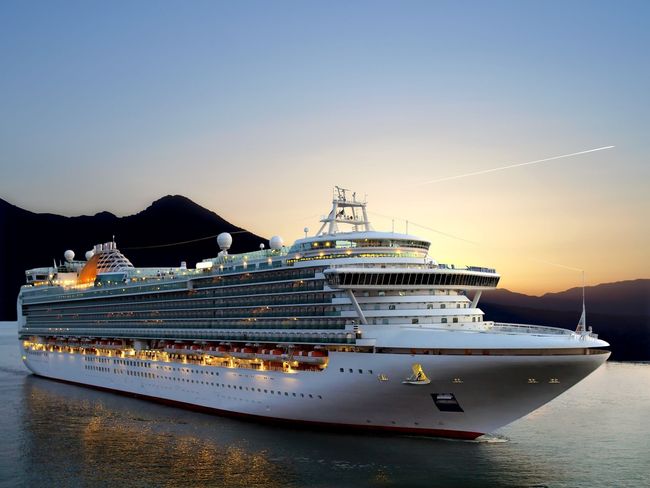 Nave da crociera che naviga su acque calme al tramonto, montagne sullo sfondo, luci accese.