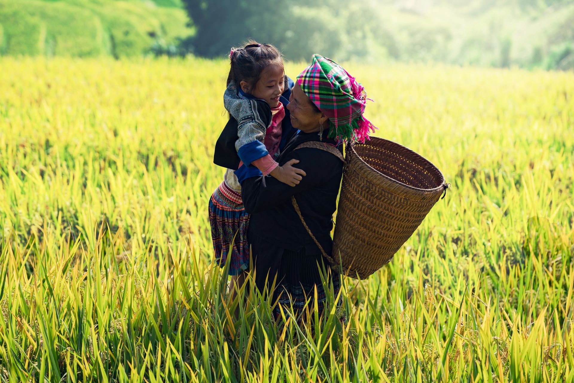 Donna con bambino in una risaia, indossa un cappello e porta un cesto; abbraccia il bambino.