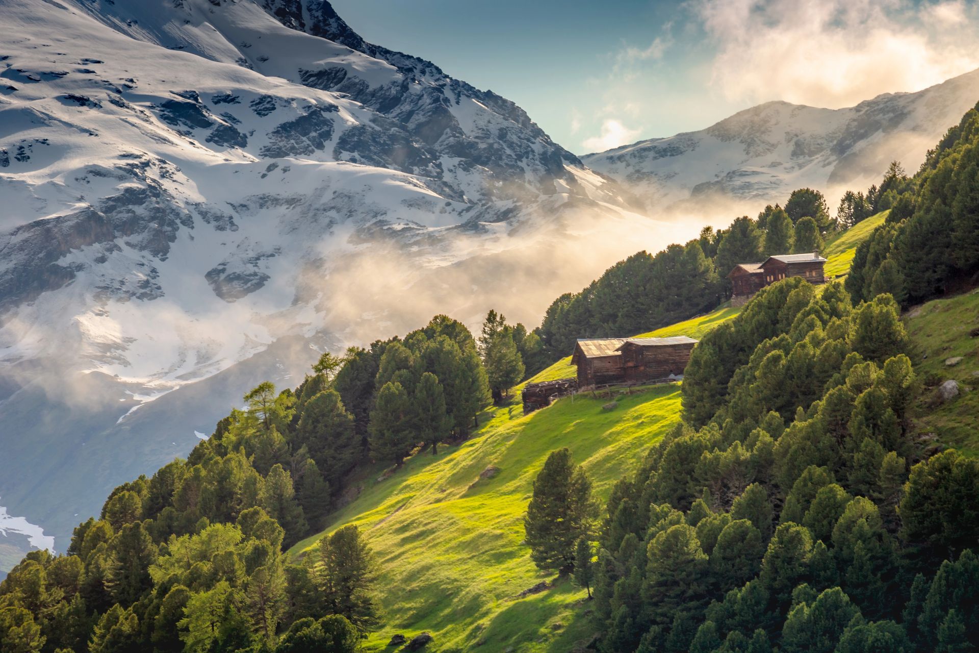 Paesaggio montano con cime innevate, pendii verdi, alberi e piccole costruzioni in legno.