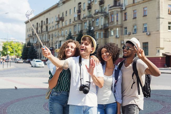 Quattro amici si scattano un selfie con un bastone in città, sorridendo e salutando.
