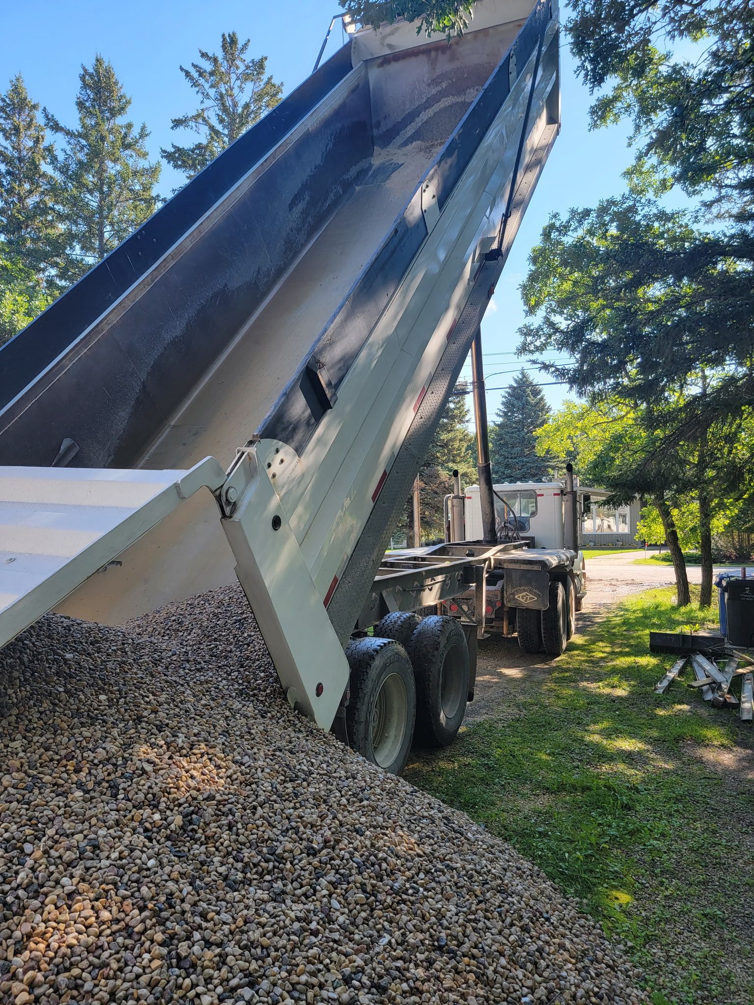 A dump truck tilted high, pouring a large pile of gravel onto the ground outdoors surrounded by green trees.