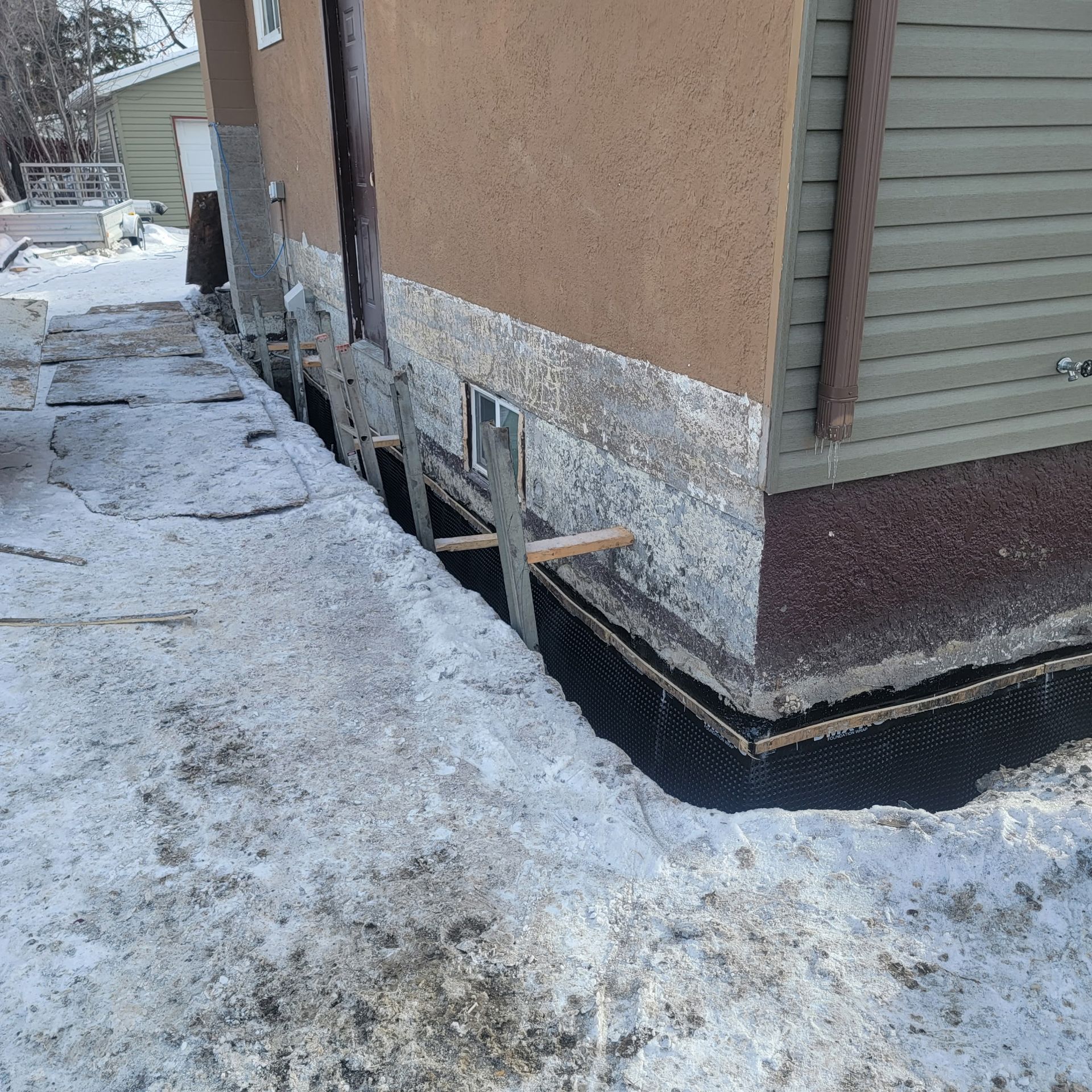 Foundation repair work on a house exterior with exposed concrete, black waterproofing membrane, and wood supports.