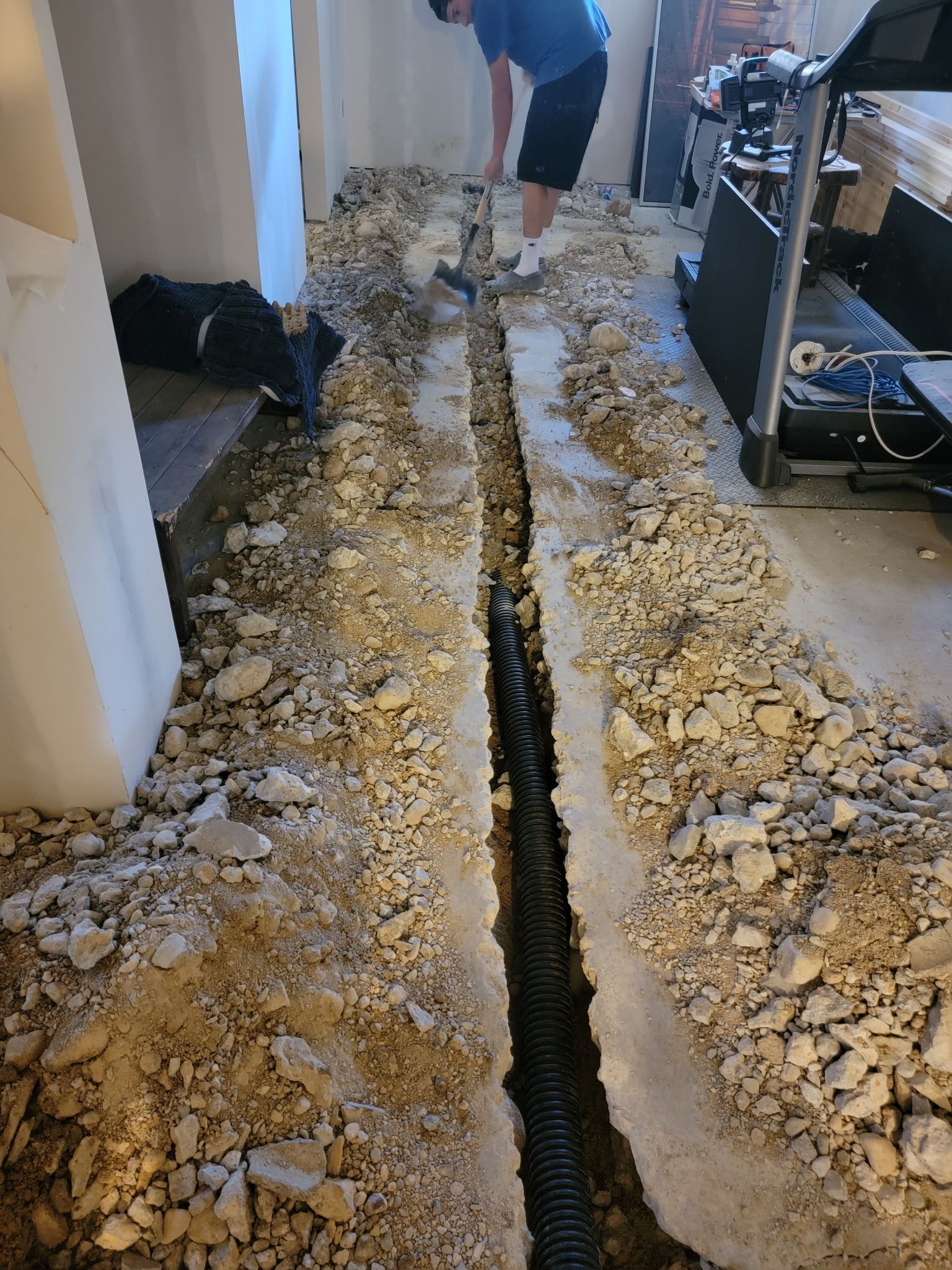 A person working in a room with a long trench cut into a concrete floor, revealing a black drainage pipe underneath.
