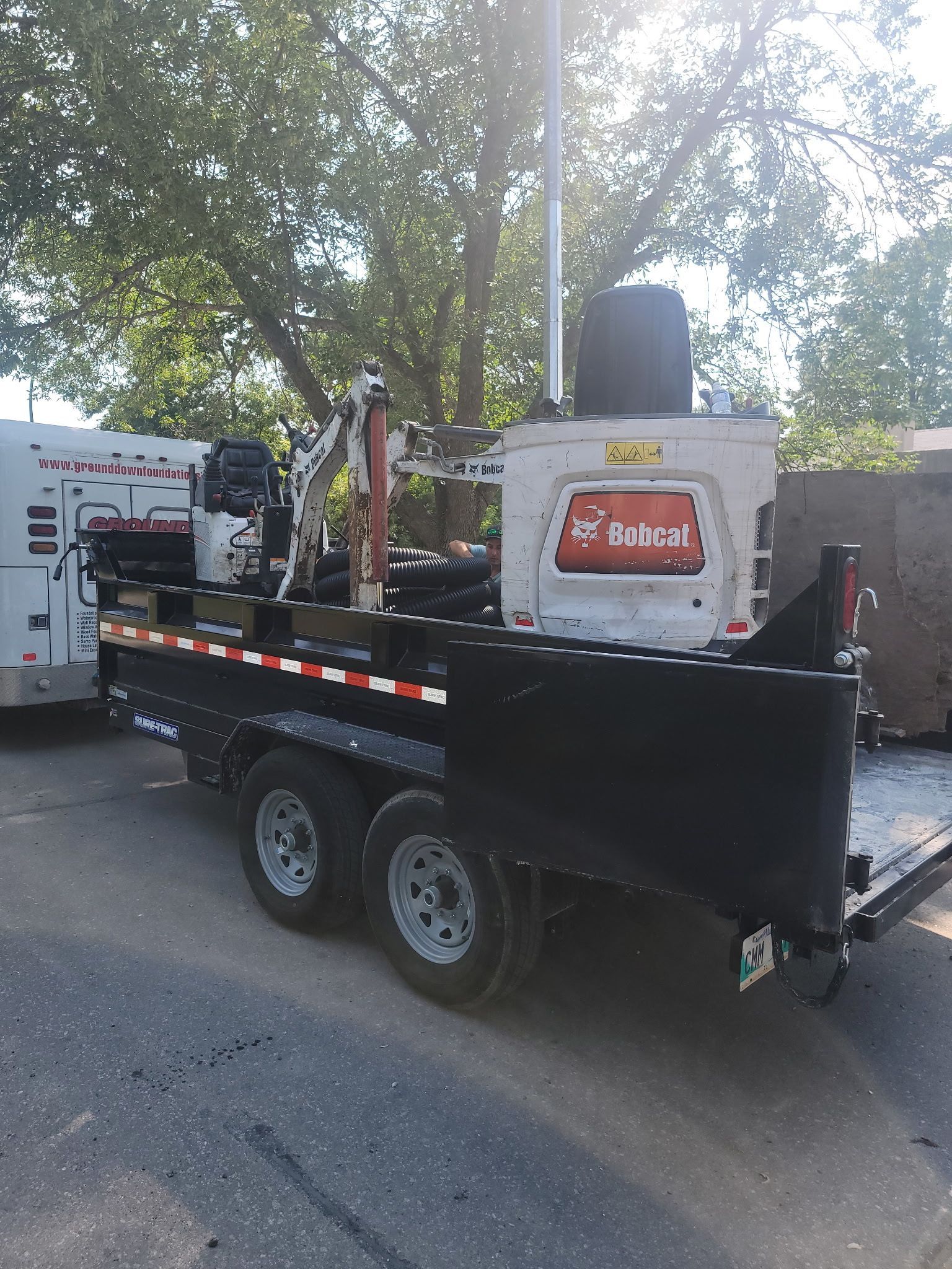A white Bobcat mini-excavator secured on a black tandem-axle utility trailer parked on an asphalt road.